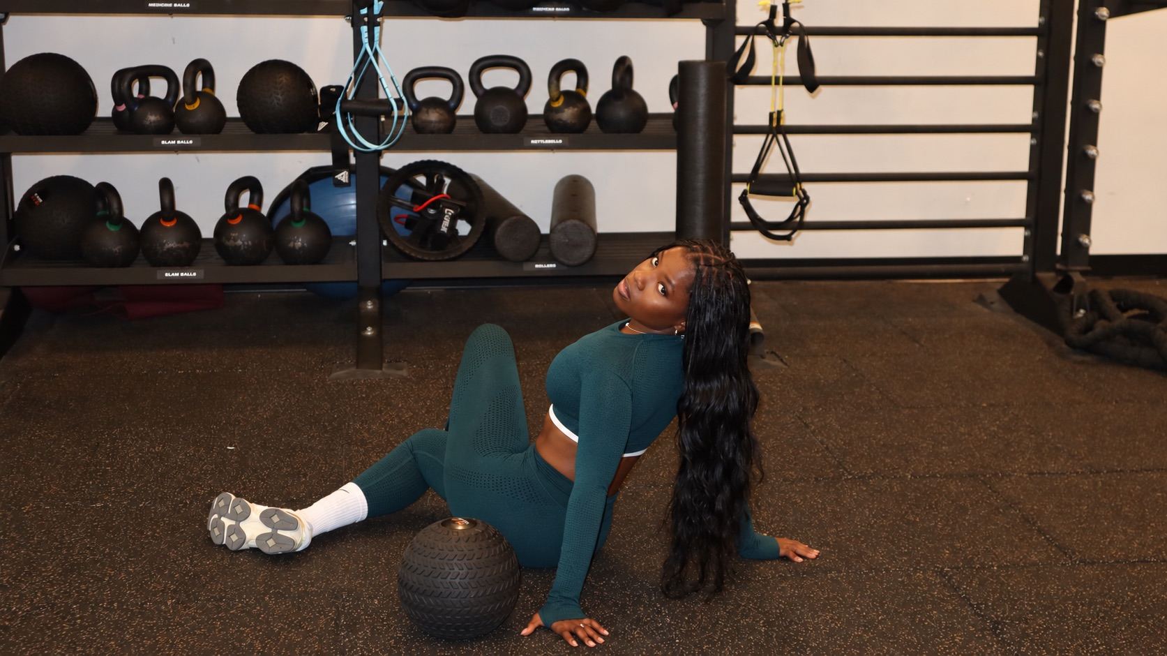 Temi sitting on the gym floor beside a slam ball, kettlebells racked behind her