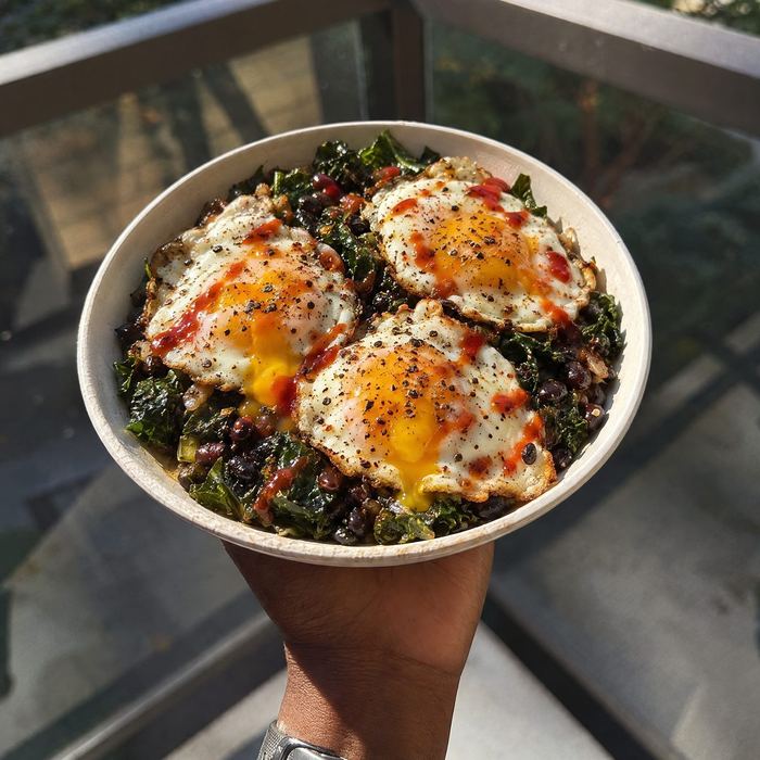 A bowl of greens and beans topped with three fried eggs and hot sauce