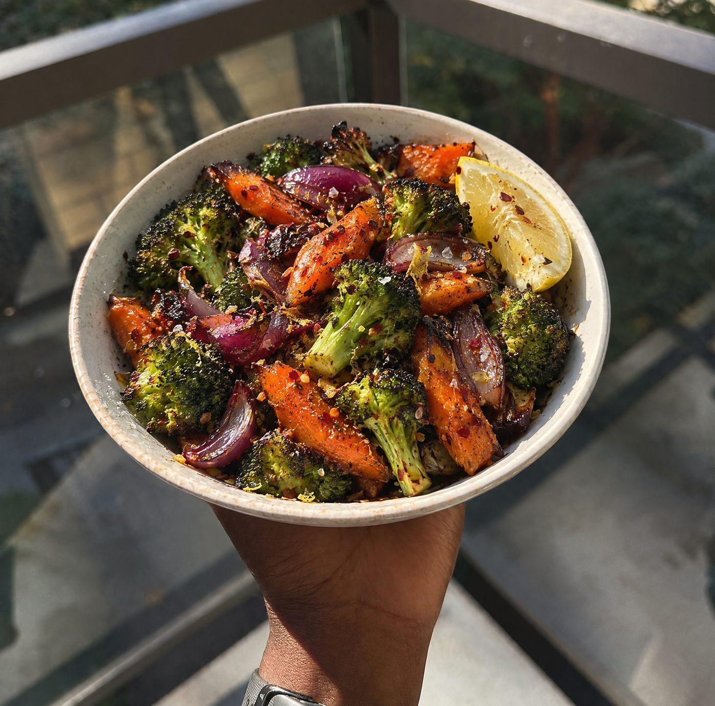 A bowl of roasted broccoli, carrots and red onion with chilli flakes and a wedge of lemon